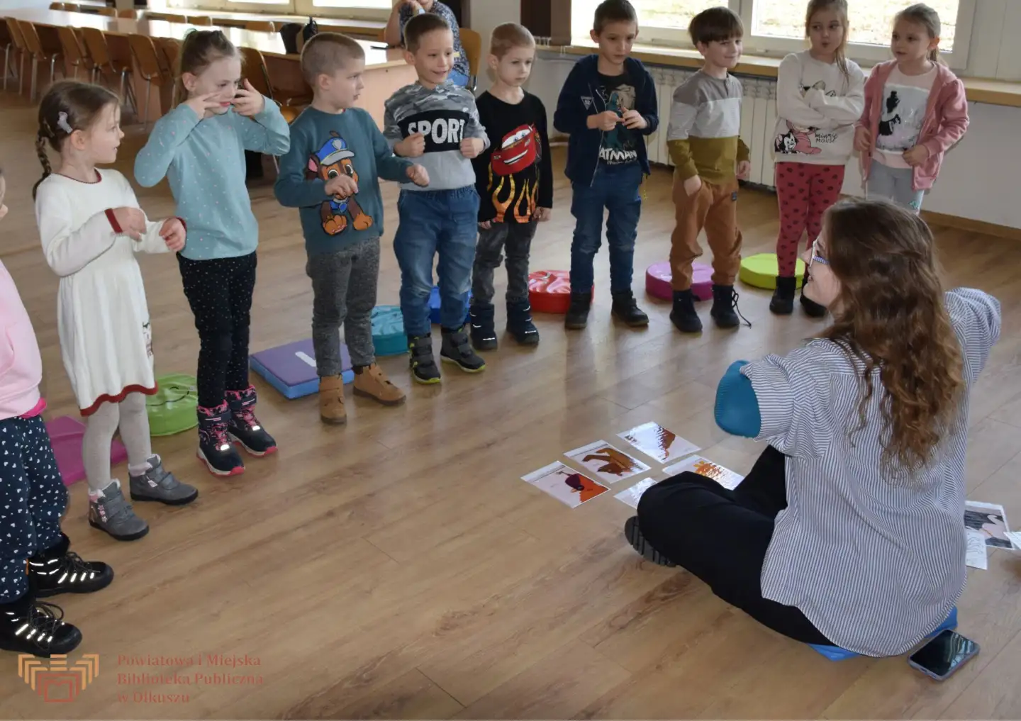 Zdjęcie zostało zrobione w trakcie Zabaw z książką po angielsku. Przedstawia grupę dzieci stojących przed siedzącą, dorosłą kobietą. Grupa pokazuje różne gesty.