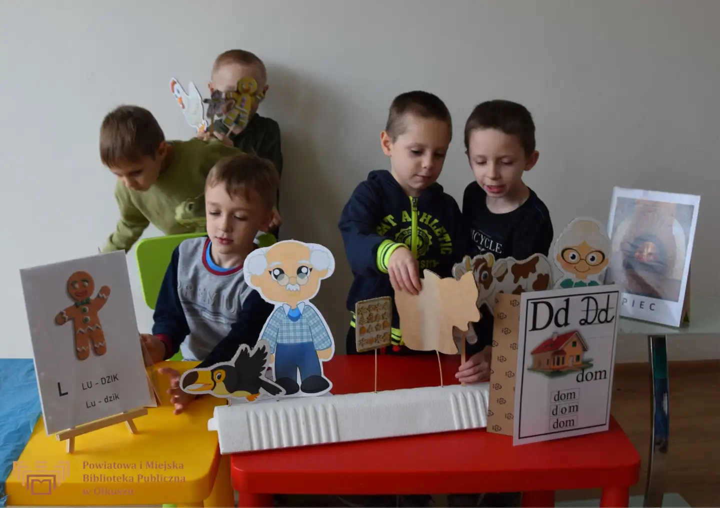 Zdjęcie zostało zrobione w trakcie Kreatywnych zabaw z książką. Grupa chłopców stoi przy stoliczkach, na którym znajdują się różne obrazki, literki i wyrazy.