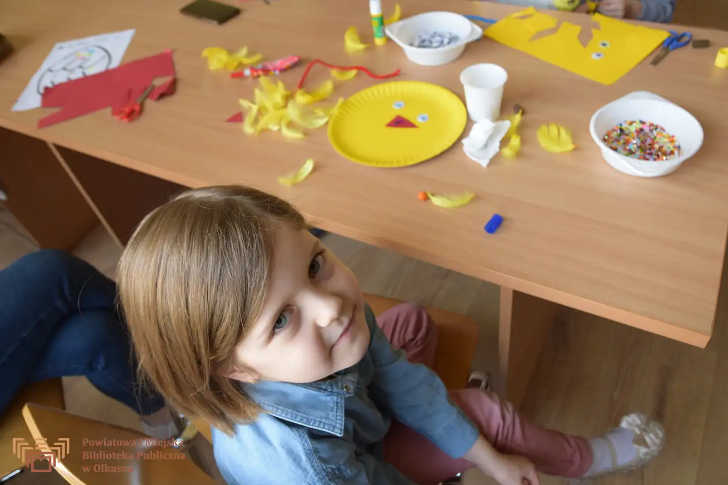 Dziewczynka siedzi na krześle i patrzy się do obiektywu aparatu. Obok niej znajduje się stolik, na którym widoczne są różne artykuły papiernicze.