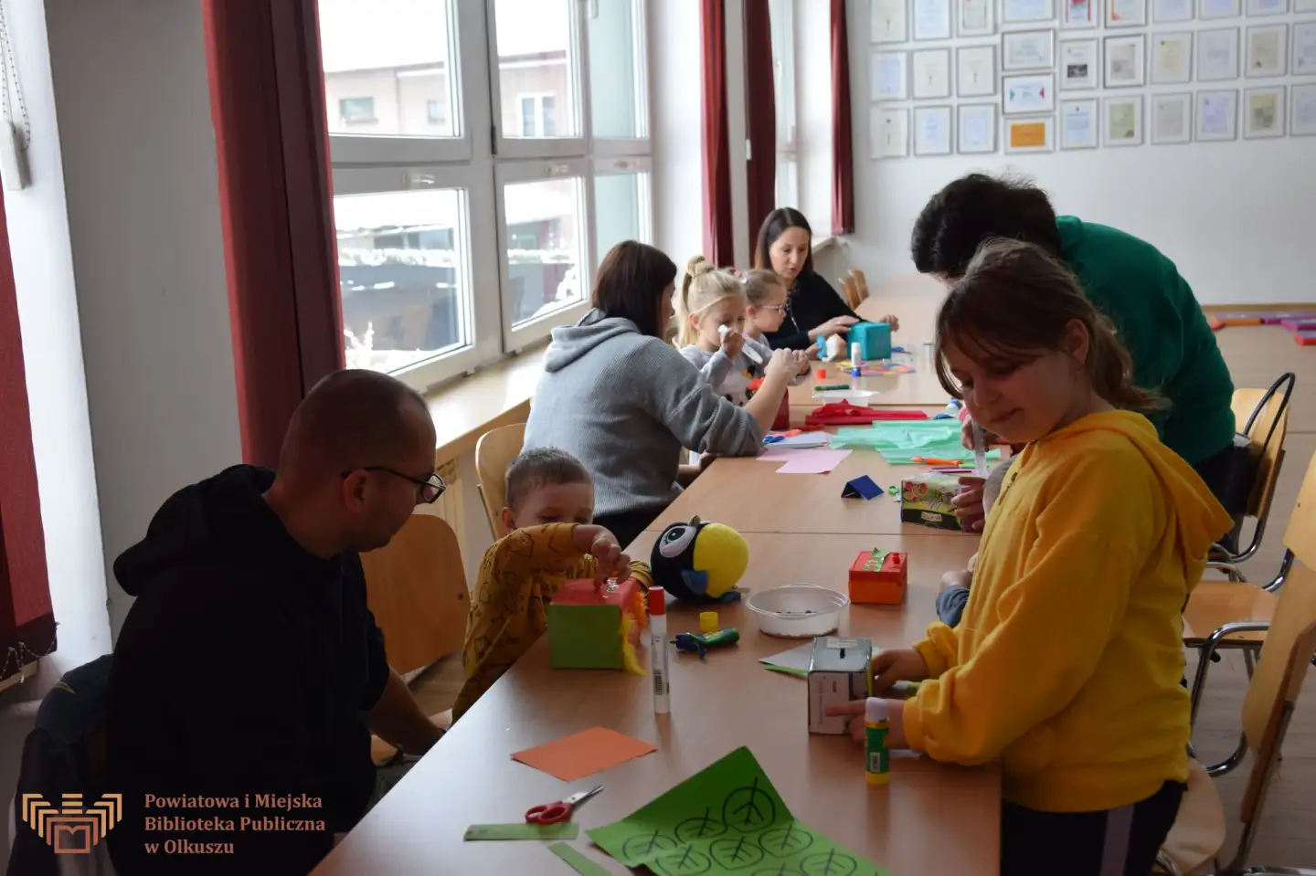 Zdjęcie przedstawia siedzące przy stoliku dzieci wraz z rodzicami.m Wspólnie wykonują kolorowe skarbonki. Na stoliku leżą kolorowe materiały plastyczne.