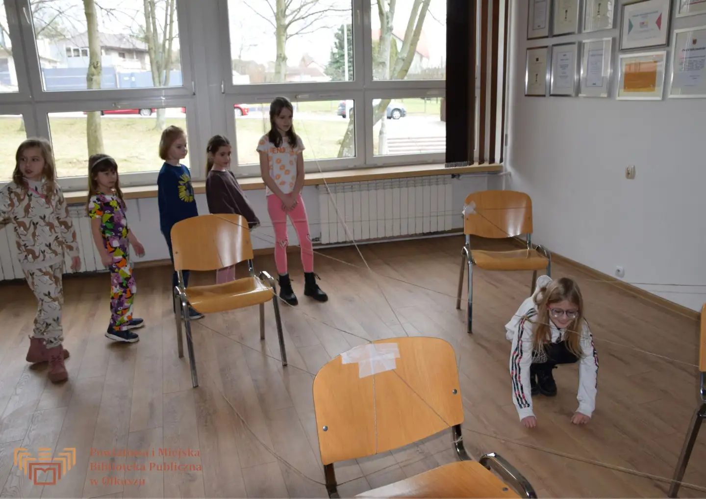Grupa dzieci stoi przy krzesełkach, między którymi przeciągnięty jest sznurek na kształt pajęczyny. Jedna dziewczynka przechodzi pod sznurkiem.