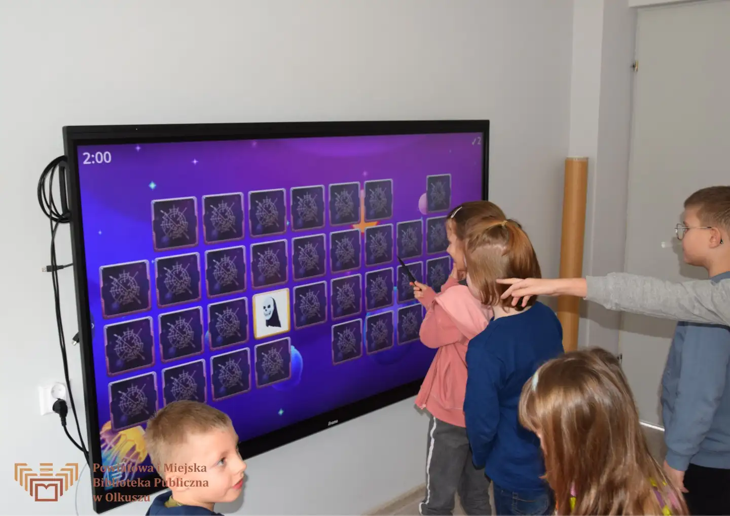 Grupa dzieci stoi przy tablicy interaktywnej. Jedna dziewczynka dotyka ekran, na którym znajdują się kafelki do gry MEMORY.