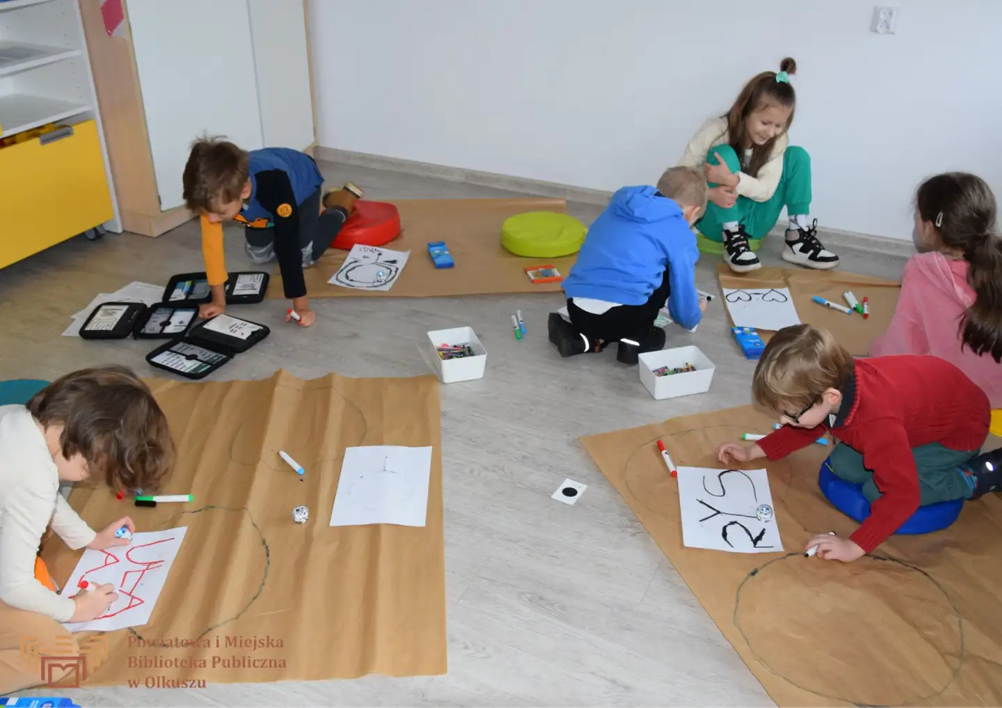 Grupa dzieci siedzi na podłodze i rysuje kształty na białych kartkach. Dorosła kobieta im pomaga. Część dzieci korzysta z małych robotów ozobotów.