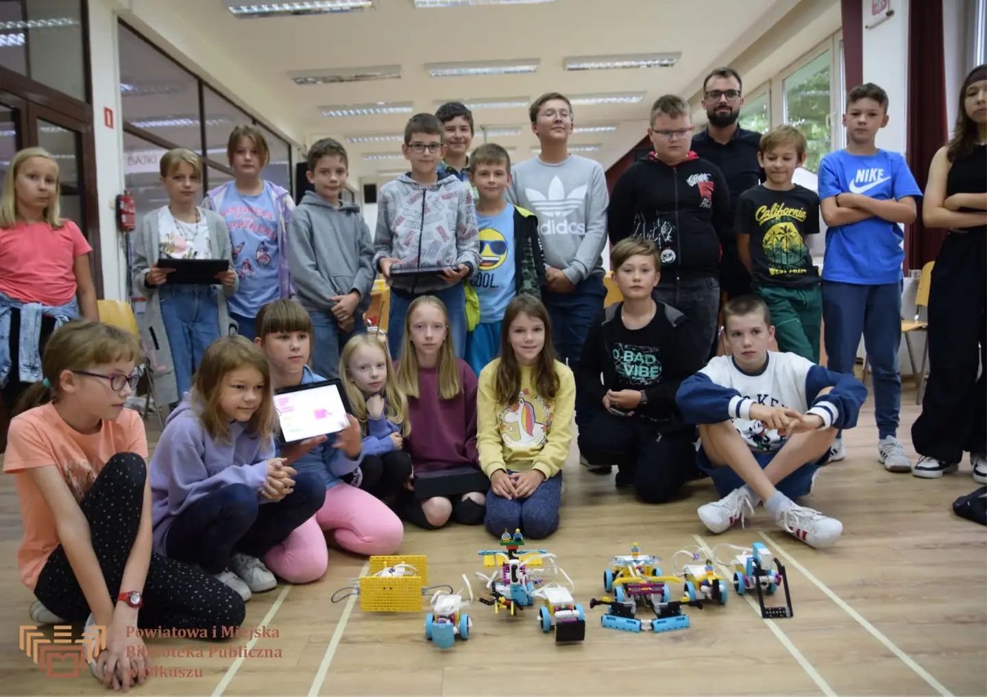 Grupa dzieci z opiekunem, część siedząca, część stojąca - wokół kilku małych robotów. Trzy osoby trzymają tablety.