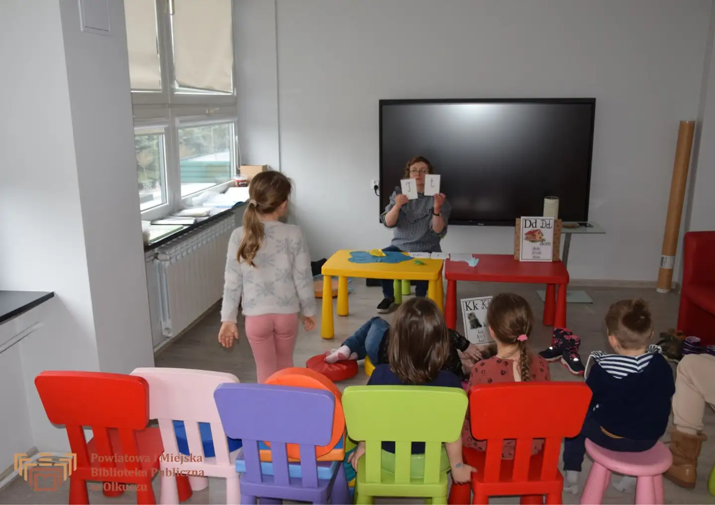 Zdjęcie zostało zrobione w salce edukacyjnej Biblioteki. Na pierwszym planie widoczne są kolorowe krzesełka i siedzące na nich dzieci. Dzieci odwrócone się w stronę Pani Prowadzącej zajęcia, która siedzi przy stoliczku i pokazuje literkę T.