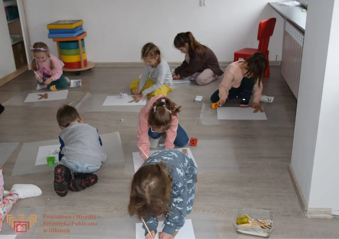 Zdjęcie zostało zrobione w salce edukacyjnej Biblioteki. Przedstawia grupę dzieci, które siedzą na podłodze i kolorują literkę K.