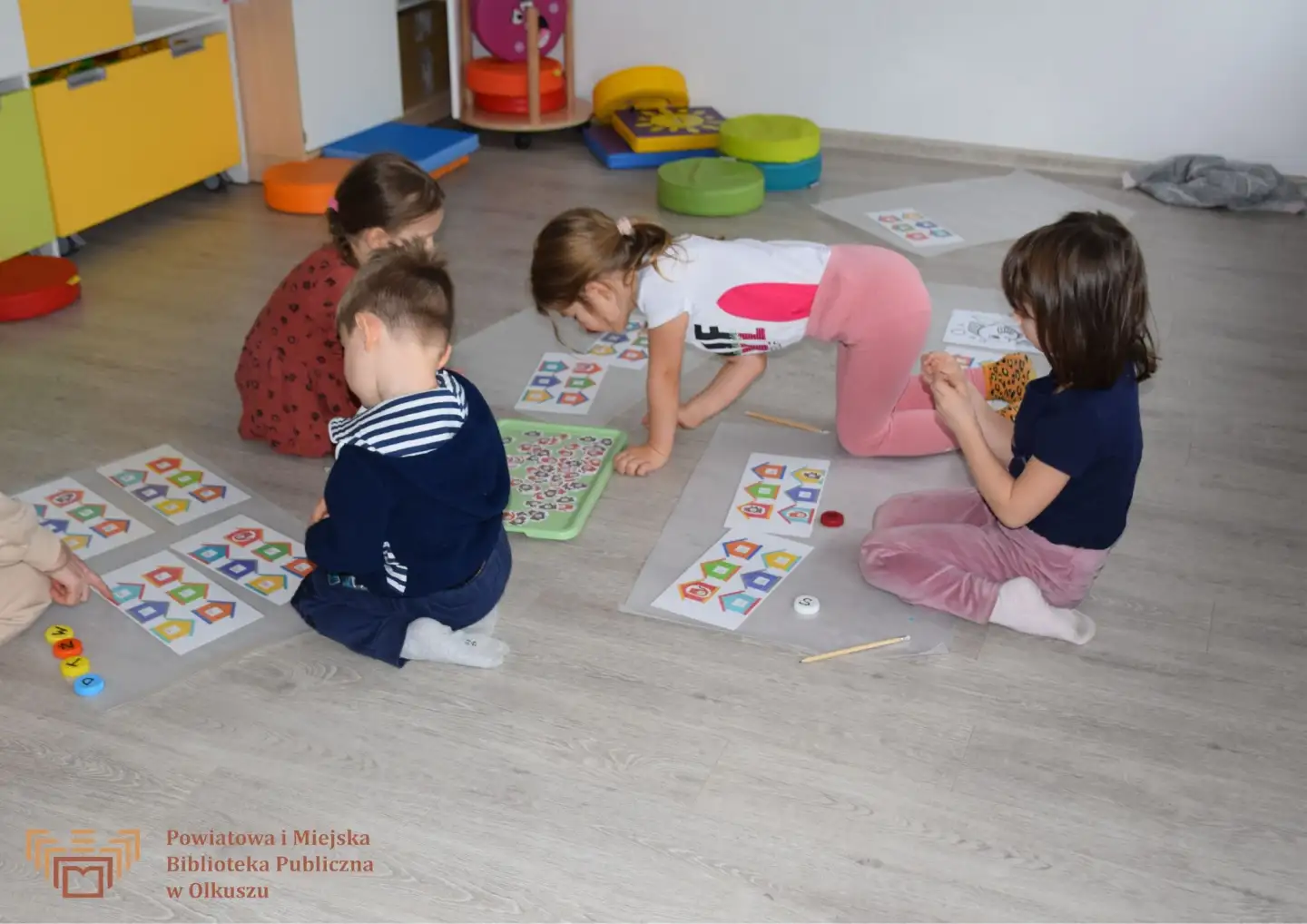 Zdjęcie zostało zrobione w salce edukacyjnej Biblioteki. Na zdjęciu widoczna jest grupa dzieci, które siedzą na podłodze i pracują z kartkami przedstawiającymi małe, kolorowe domki.