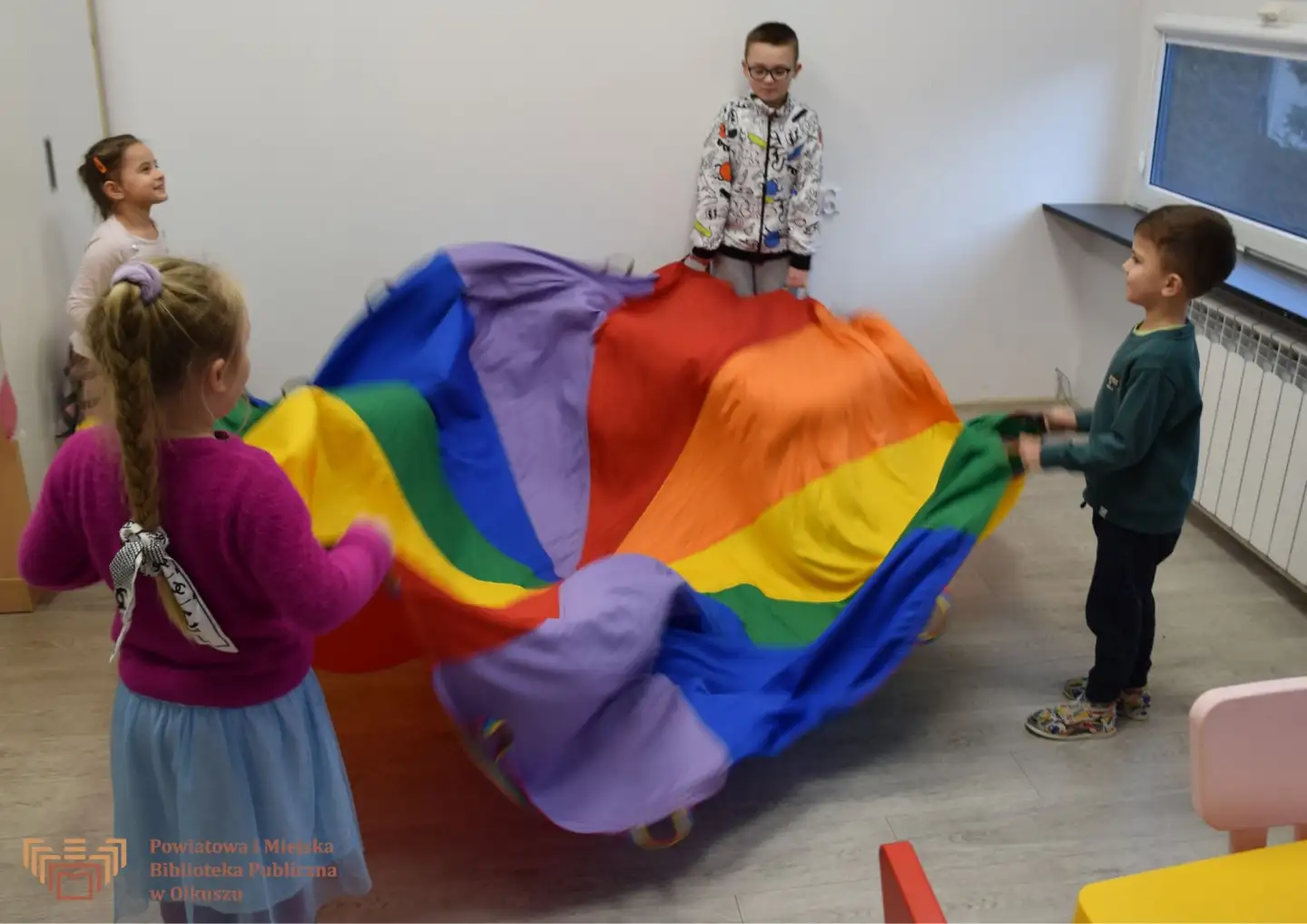 Zdjęcie zrobione w trakcie zajęć Zabawy z książką po angielsku. Czwórka dzieci unosi kolorową chustę.
