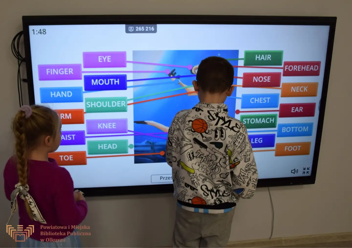 Zdjęcie zrobione w trakcie zajęć Zabawy z książką po angielsku. Dziewczynka z chłopcem stoją przy tablicy interaktywnej. Na ekranie widoczne nazwy części ciała po angielsku.