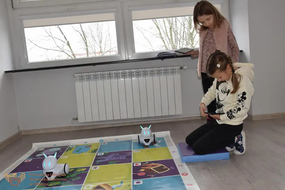 Zdjęcie przedstawia dwie dziewczynki z tabletami w rękach. Za ich pomocą kierują robotami - photonami jeżdżącymi po specjalnej, kolorowej macie.