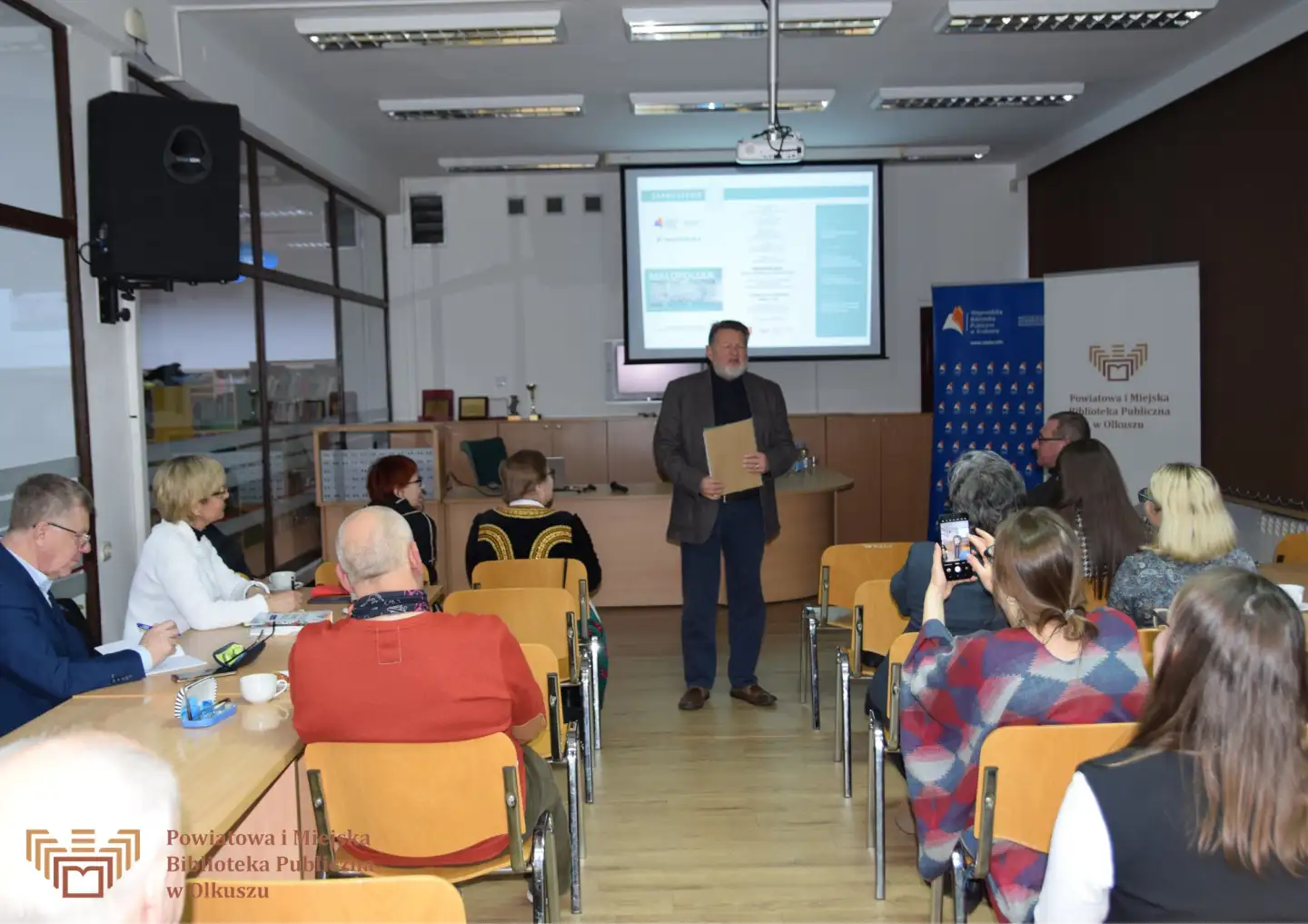 Dorosły mężczyzna przemawia do zgromadzonych osób, siedzących na krzesłach. W tle widać szyld PiMBP w Olkuszu, Wojewódzkiej Biblioteki Publicznej w Krakowie oraz ekran projektora.