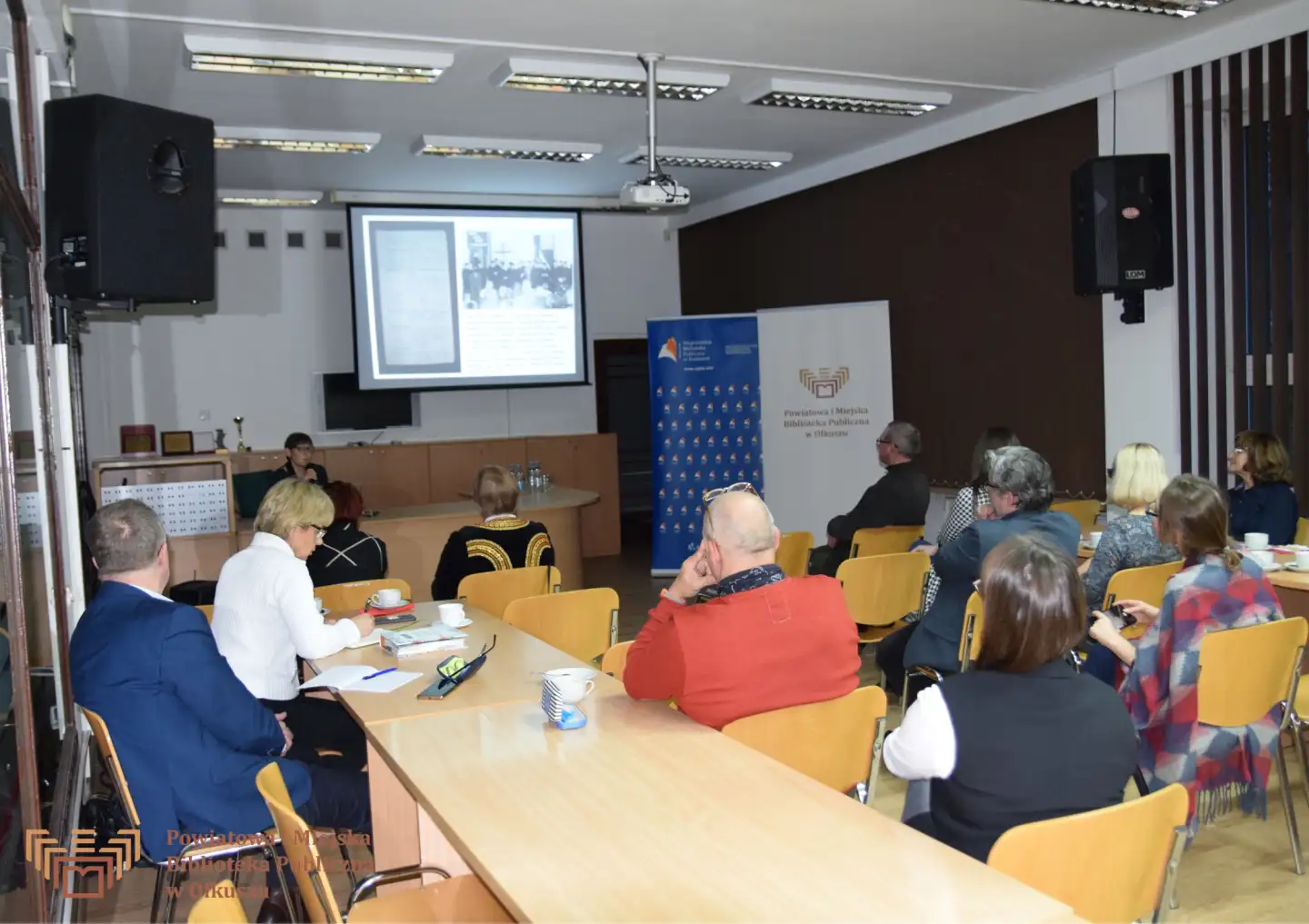 Dorosła kobieta siedzi za biurkiem i przemawia do zgromadzonych osób, siedzących na krzesełkach. W tle widać szyld PiMBP w Olkuszu, Wojewódzkiej Biblioteki Publicznej w Krakowie oraz ekran projektora.