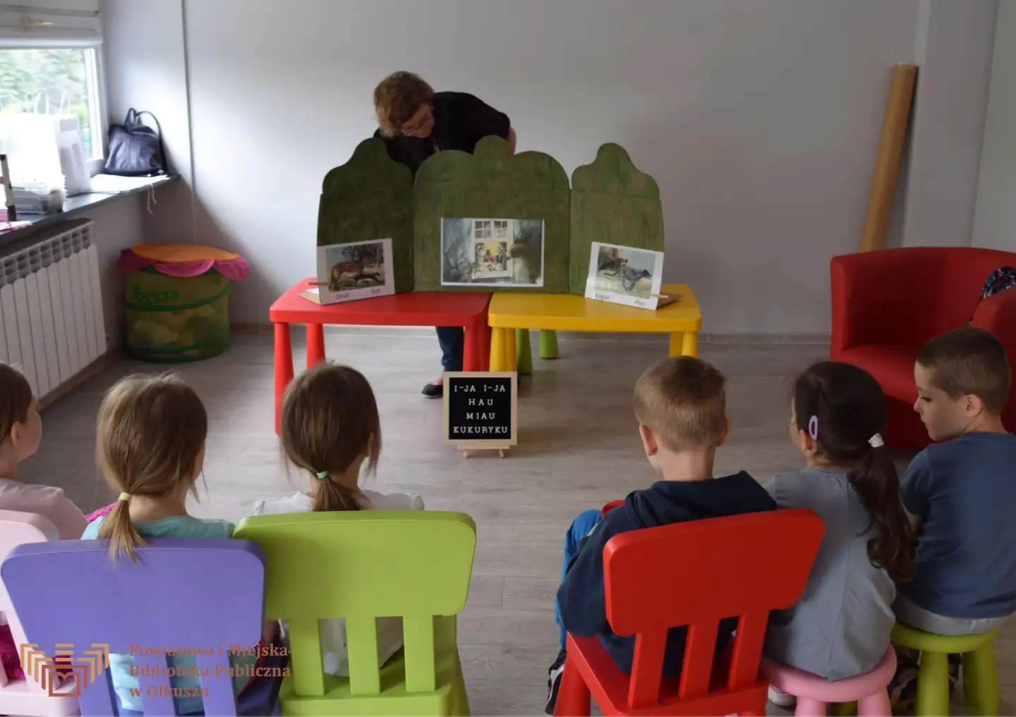 Na pierwszym planie grupa dzieci siedząca na krzesłkach. Na drugim planie dwa stoliczki z obrazkami z książek oraz Pani pochylająca się nad ilustracjami.