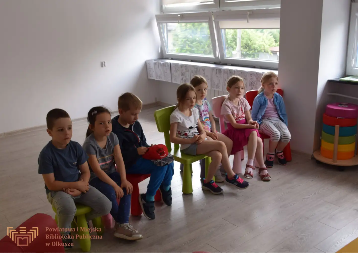 Grupa dzieci siedząca na krzesełkach, słuchająca Pani prowadzącej zajęcia