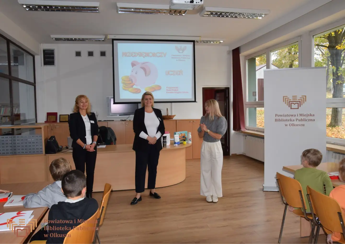 Zdjęcie zostało zrobione w Czytelni olkuskiej Biblioteki. Przedstawia trzy Panie prowadzące wydarzenie - w tym dwie reprezentujące bank PEKEO S.A. Panie stoją po środku zdjęcia, za nimi znajduje się ekran z prezentacją, na której widnieje tekst: PRZEDSIĘBIORCZY UCZEŃ oraz zdjęcie świnki skarbonki z monetami.