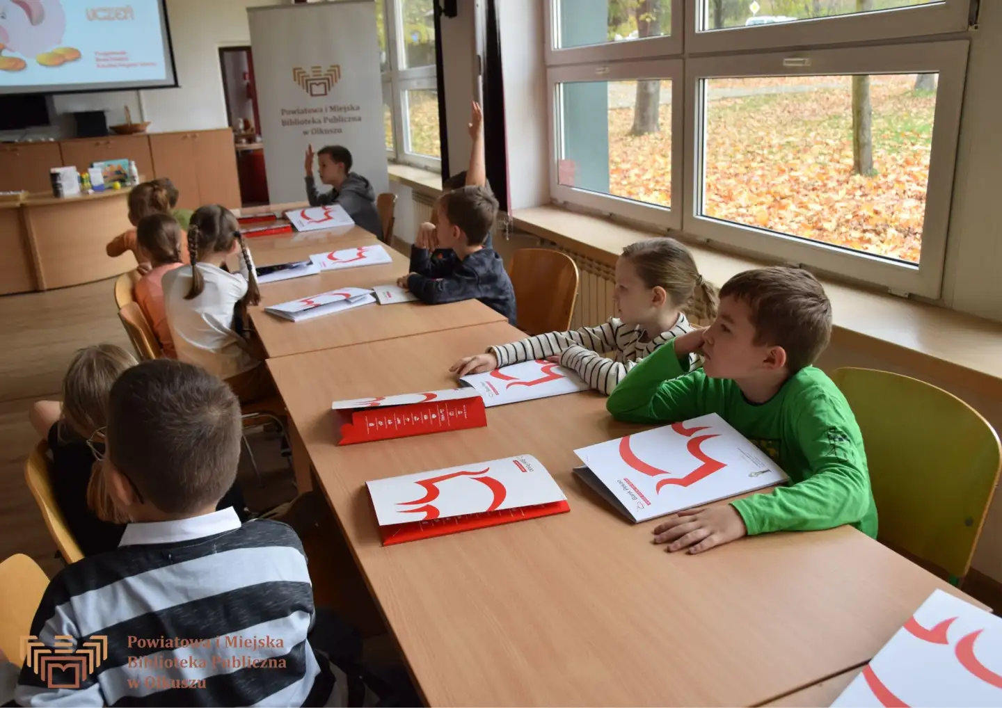 Zdjęcie zostało zrobione w Czytelni olkuskiej Biblioteki. Przedstawia dzieci siedzące przy stoliku. Przed nimi leżą biało-czerwone teczki z logo Panku PKO S.A.