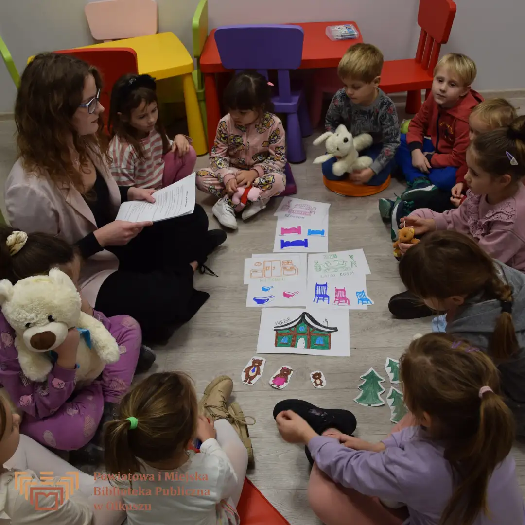 Zdjęcie zostało zrobione w Bibliotece. Przedstawia grupę dzieci siedzących na podłodze w kole. Pośrodku znajdują się wycinanki przedstawiające elementy bajki ,,Złotowłosa i trzy misie". Koło dzieci siedzi Pani prowadząca, trzymająca białe kartki z tekstem.