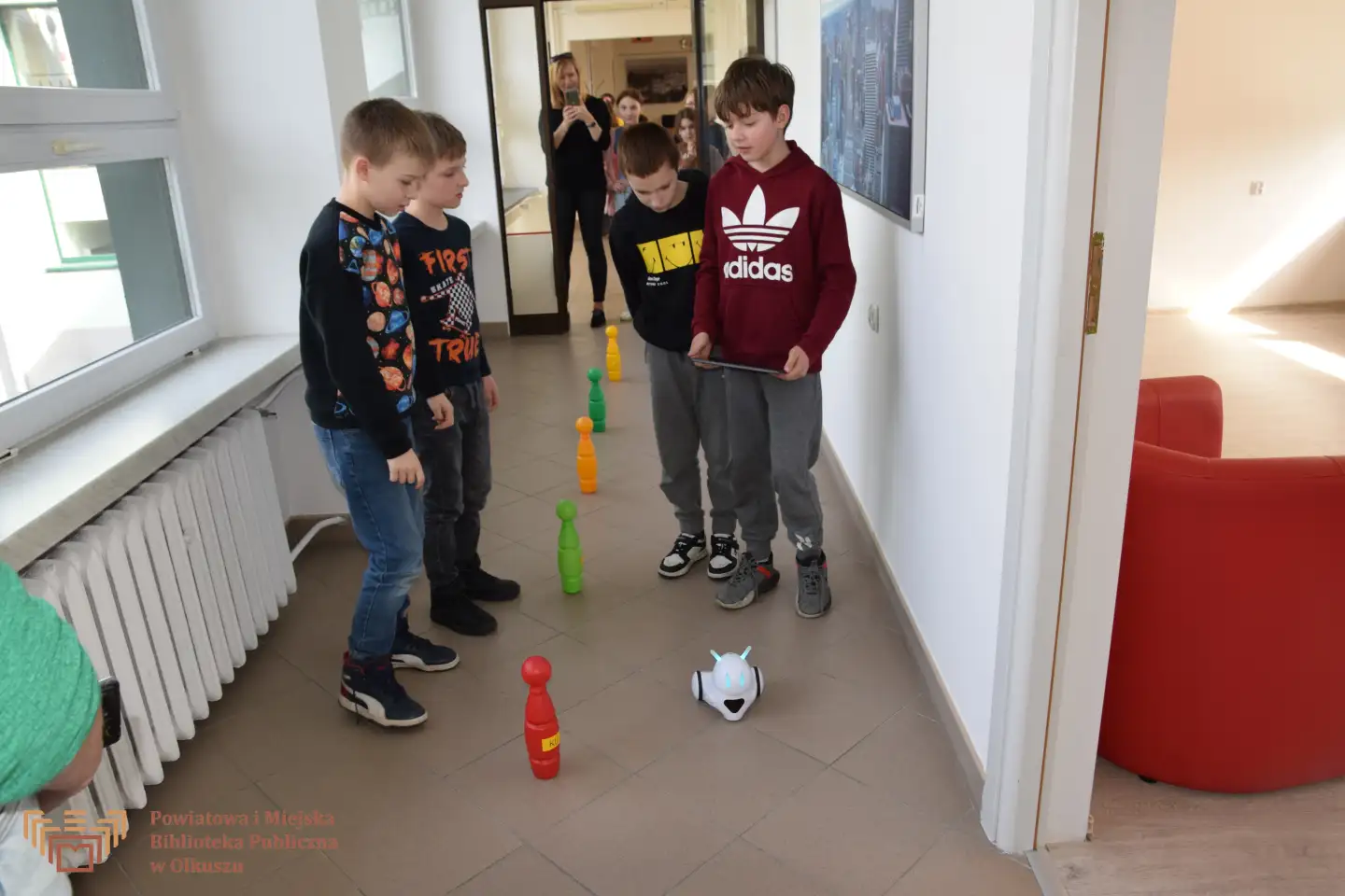 Grupa chłopców stoi w korytarzu i patrzy na robota, który jedzie koło pionów do kręgli ułożonych wzdłuż linii.